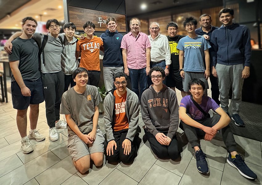The UT Programming Team gathered together for a photo in a restuarant. Eight students and three faculty coaches stand in the back row. Four students sit in the front row.