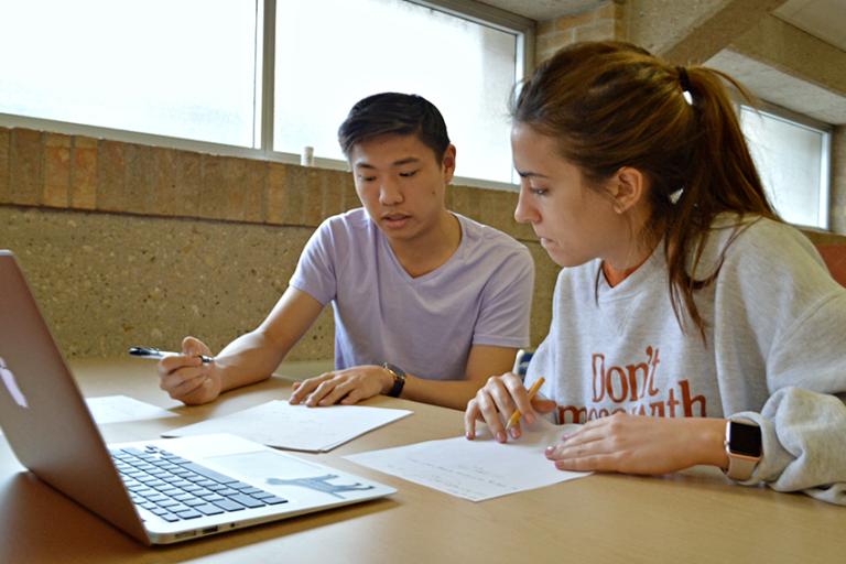 Two students work together at a table with notebooks and a laptop, receiving tutoring or study support.