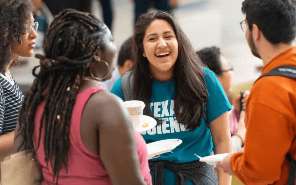 Group of students standing together indoors, talking and holding food plates
