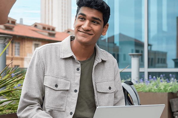 A student with their laptop open is smiling while sitting outside with buildings in the background.