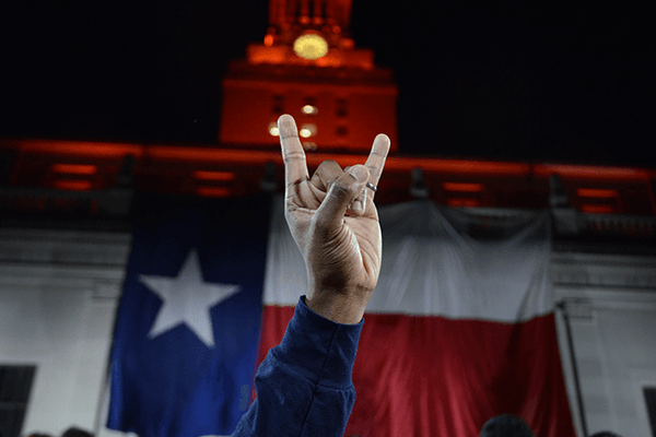 A hand makes the “Hook ’em” gesture in front of the Texas flag and orange-lit UT tower at night.