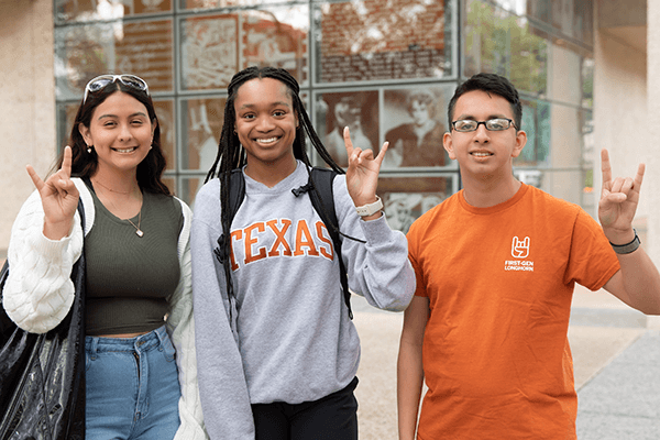 Three students are standing outdoors, smiling and making the "Hook 'em" hand gesture