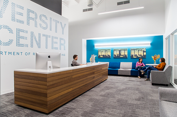 A modern reception area with a wooden desk, a person sitting behind it, and two people seated and talking in a lounge with blue accent walls.