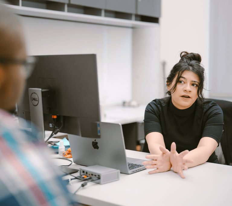Two people have a conversation in an office; one sits at a computer facing the other across the desk in a professional setting.