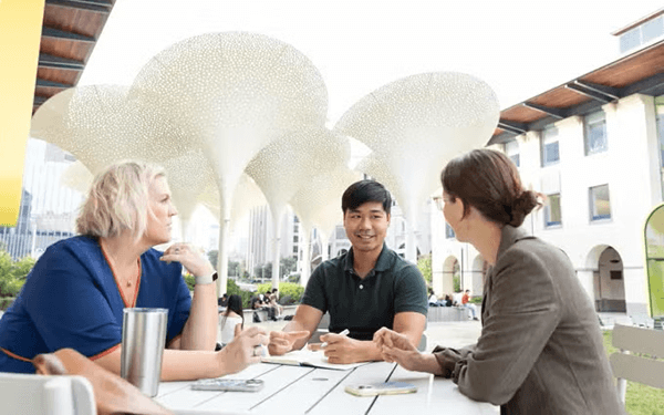 Three people sit around a white outdoor table having a discussion, with unique, large sculptural canopies and a campus building in the background.