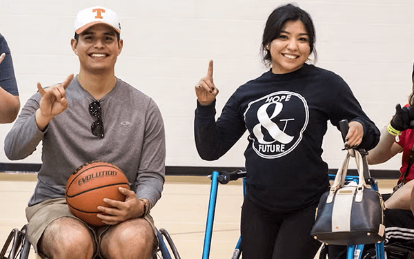 People in wheelchairs and standing, engaged in a basketball activity, showing Hook 'em hand signs and holding a basketball indoors