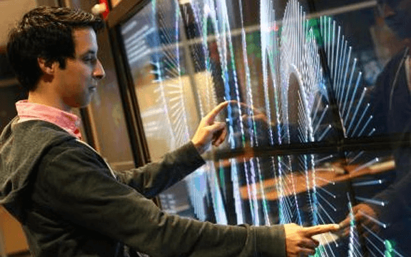 A person uses both hands to interact with a large wall-mounted touchscreen displaying dynamic, multicolored data visualizations and wave-like graphs in a modern, technology-focused environment.
