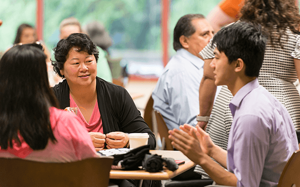 Several students and faculty are seated at a table having a conversation in a busy indoor setting, with papers, a cup, and other small items on the table.