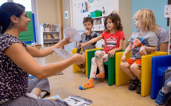 An adult sits on the floor in a classroom showing paper cutouts to a group of young children who are seated on colorful cube chairs, with bookshelves and classroom materials visible in the background.
