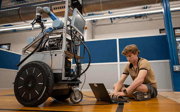 A person sits cross-legged on a gymnasium floor operating a laptop, which is wired to a large mobile robot featuring a robotic arm, two large wheels, and multiple electronic components and displays, in an industrial or laboratory setting.