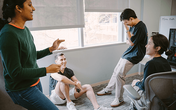 Four students in a casual discussion inside a small, well-lit office with large windows