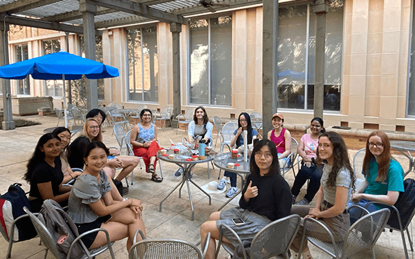 A group of students sitting around tables outdoors, engaging in conversation at a patio area on campus
