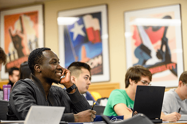A smiling student uses a notebook and pen, while in the background other students with laptops are visible, all focused on the class. 