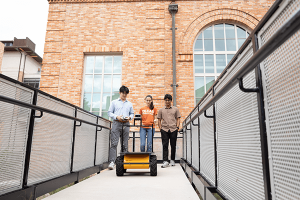 Three people standing on a walkway next to a building with a small yellow robotic vehicle positioned in front of them