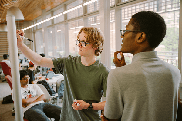 Two people are interacting in a study space, with one person holding a whiteboard marker and talking to the other.