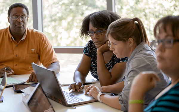 Four people sitting around a table in a brightly-lit room, focused and working together on laptops and papers
