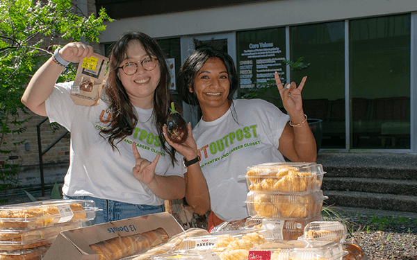 Two people in “Outpost” t-shirts pose outdoors with packaged pastries and canned drinks on tables.