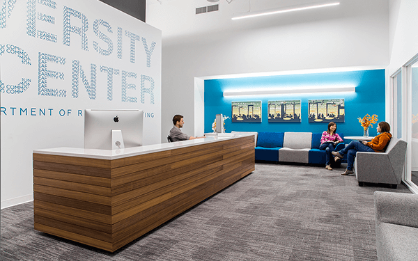 A modern reception area with a wooden desk, a person sitting behind it, and two people seated and talking in a lounge with blue accent walls