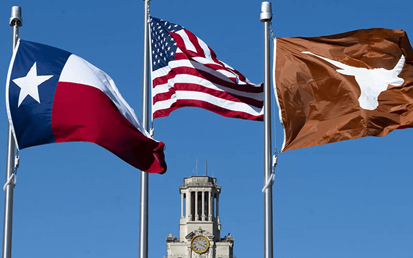 Three flags, the Texas state flag, the United States flag, and the University of Texas Longhorn flag, fly in front of a clock tower under a clear blue sky.