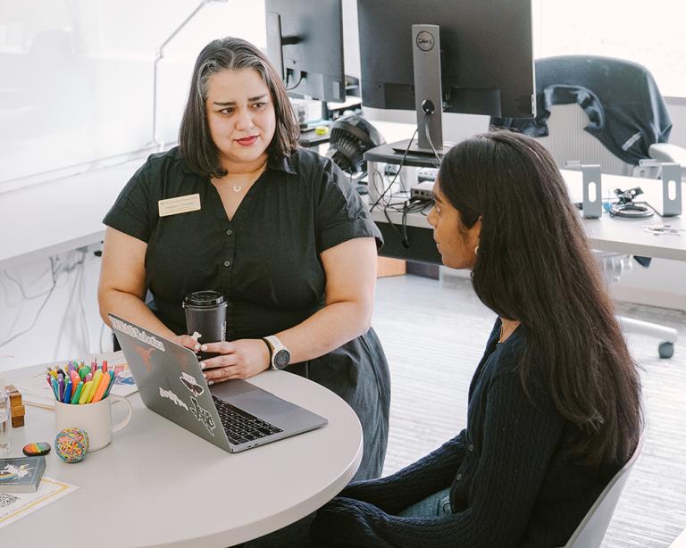 Two people are sitting and talking in a student support setting, with one person wearing a name tag and appearing to offer guidance to the other.