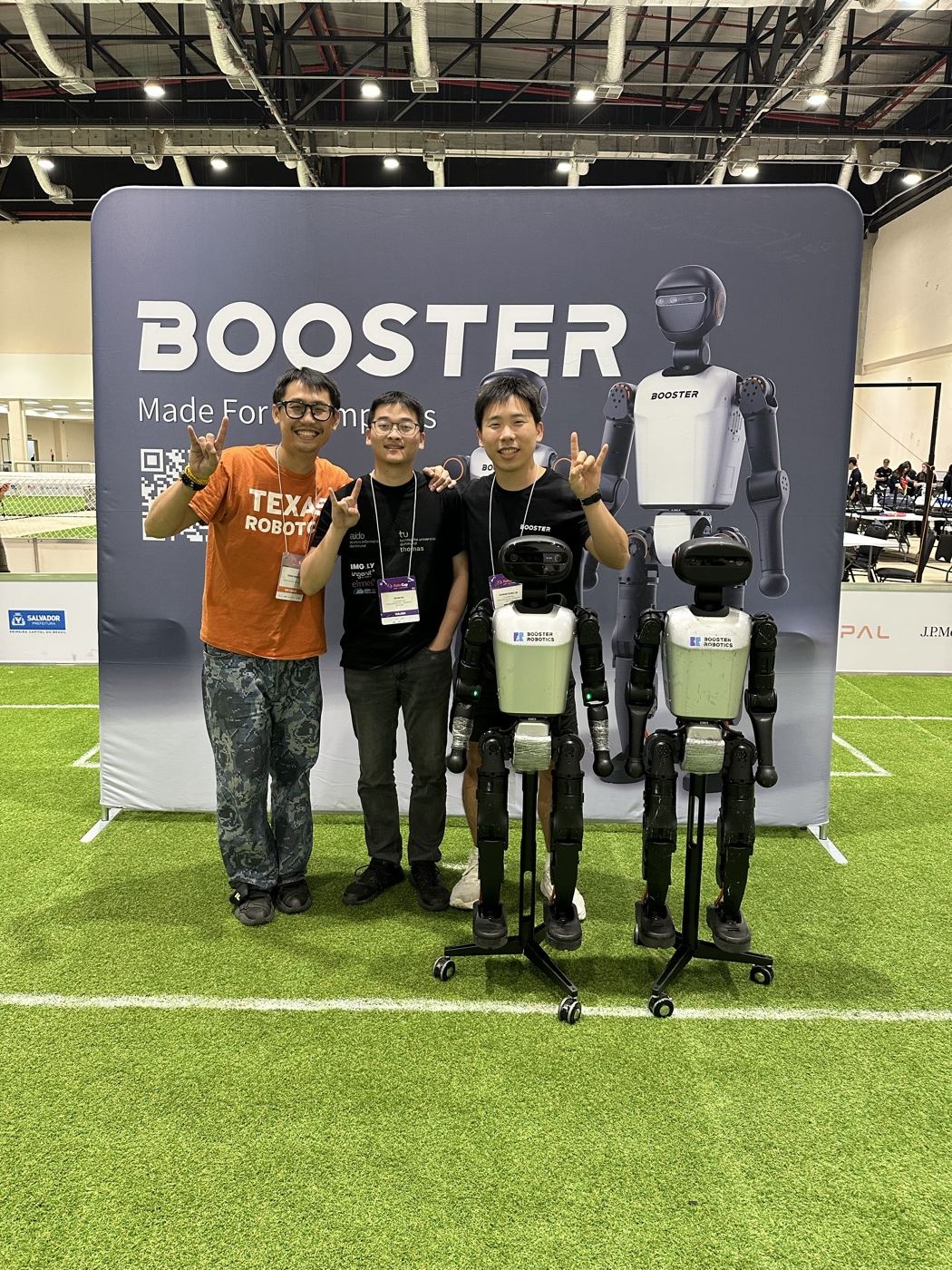 Three UT Austin Villa team members with two Booster T1 robots in front of a Booster Robotics backdrop at RoboCup 2025 in Salvador