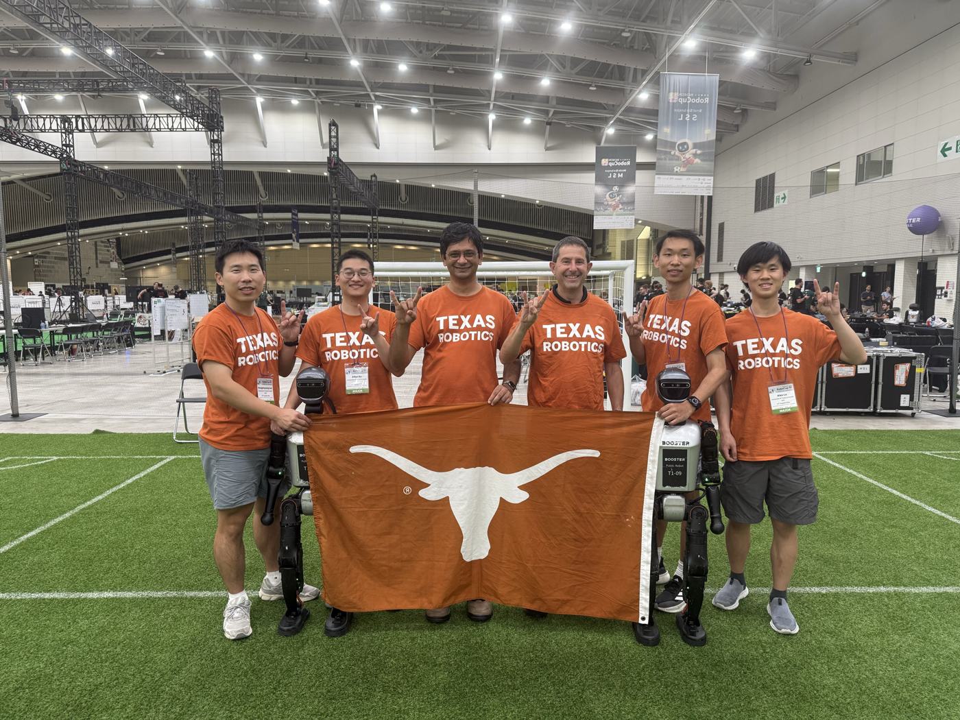 The 2026 UT Austin Villa team in Texas Robotics shirts, holding a Longhorn flag beside two Booster T1 robots on an indoor soccer field